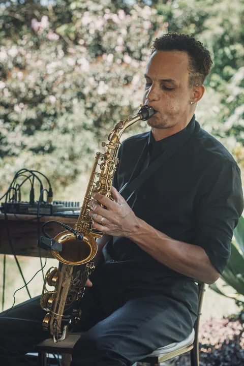 Saxofonista de camisa preta tocando ao ar livre, ao lado de equipamento de som