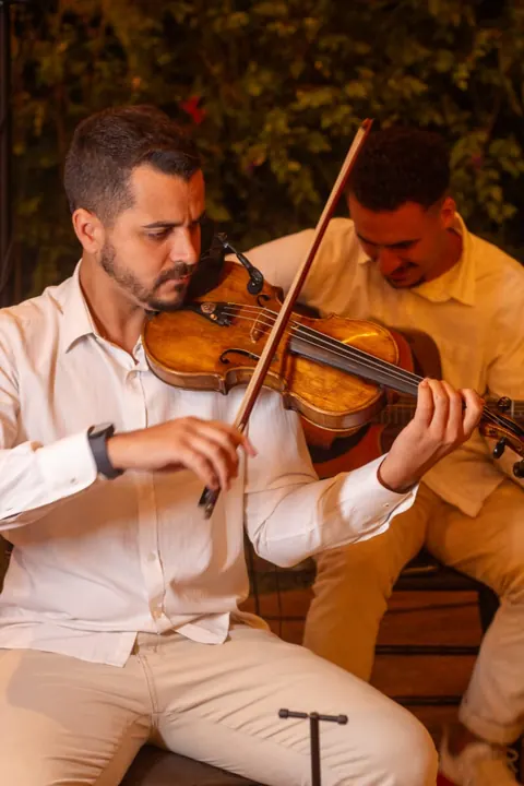 Violinista de camisa branca tocando ao ar livre, com outro músico ao fundo