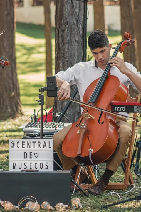 Violoncelista tocando ao ar livre ao lado de uma placa com o Instagram da Central de Músicos e equipamento de som